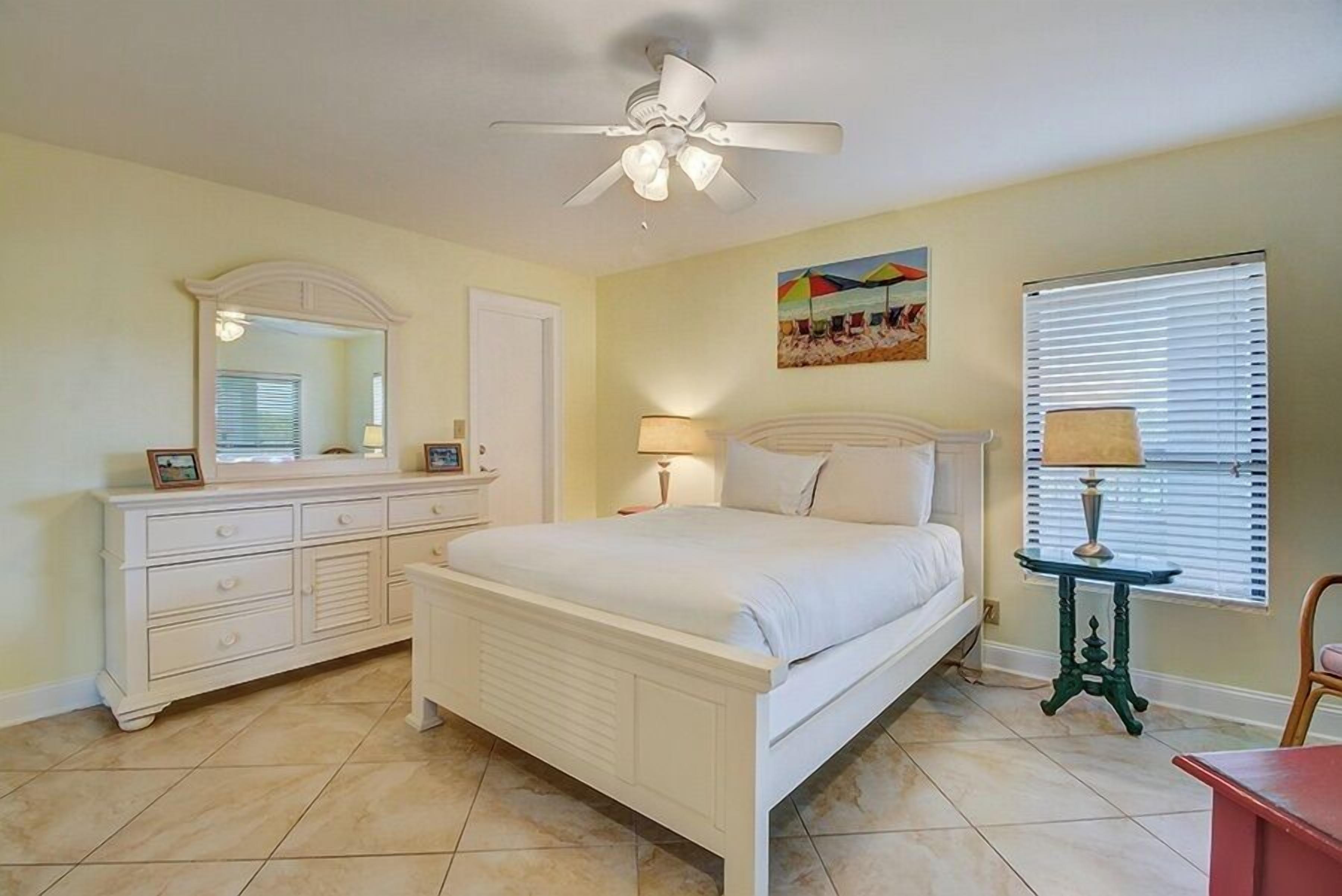 A bedroom with a bed, a dresser, a mirror, a ceiling fan, a window with blinds, and a painting on the wall.
