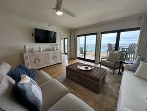 A living room with a large flat-screen TV, a white cabinet, a wicker coffee table, a sofa, and a chair.
