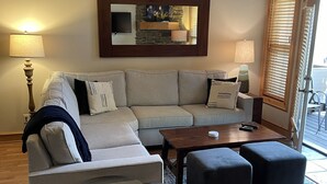 Un soggiorno con un divano color beige, due poggiapiedi, un tavolino da caffè e una TV montata a parete.