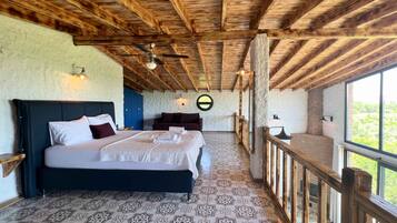 Quarto com cama grande, vigas de madeira no teto, ladrilhos com padrão no chão, vista através de uma varanda e decoração circular na parede.