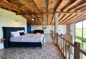 Quarto com cama grande, vigas de madeira no teto, ladrilhos com padrão no chão, vista através de uma varanda e decoração circular na parede.