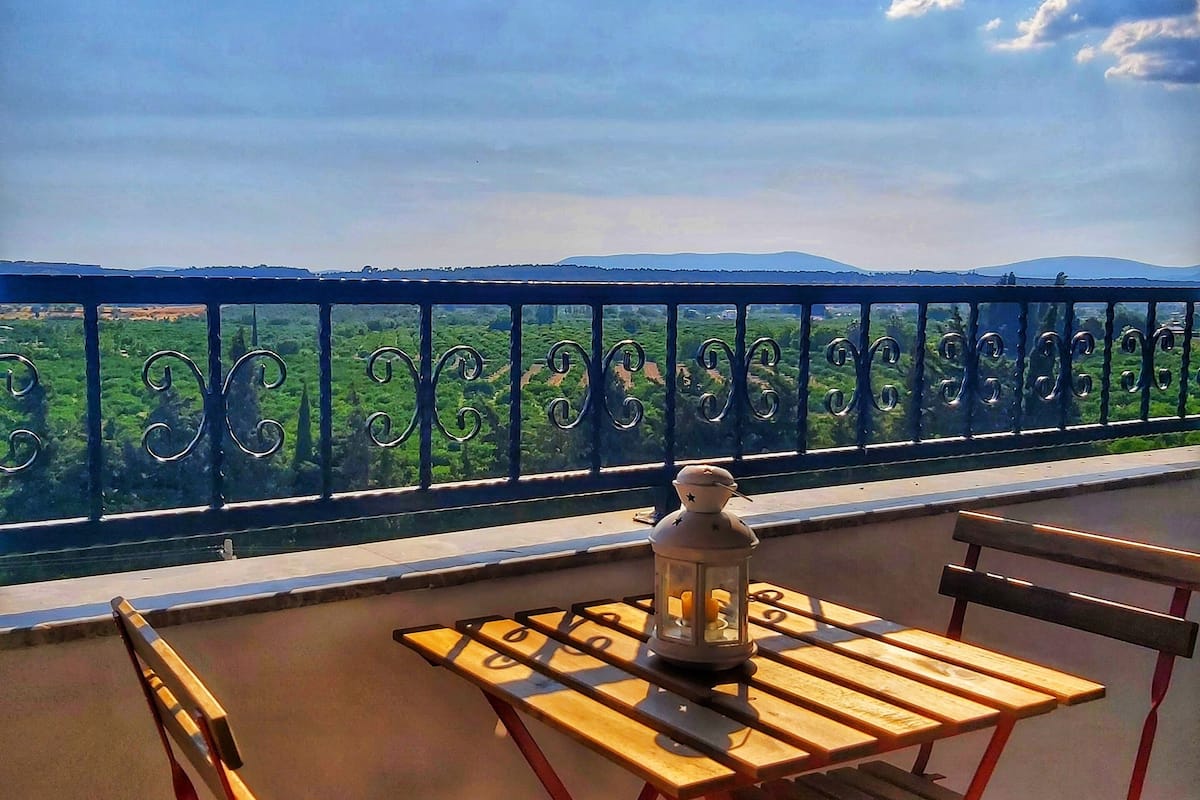 Uma varanda com mesa e cadeiras, um candeeiro e vista para uma paisagem verde com colinas.