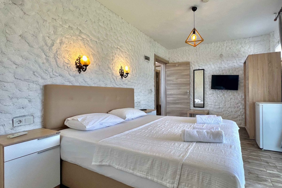 Quarto com uma cama grande, mesas de cabeceira, guarda-roupa, televisão e luminária pendente.