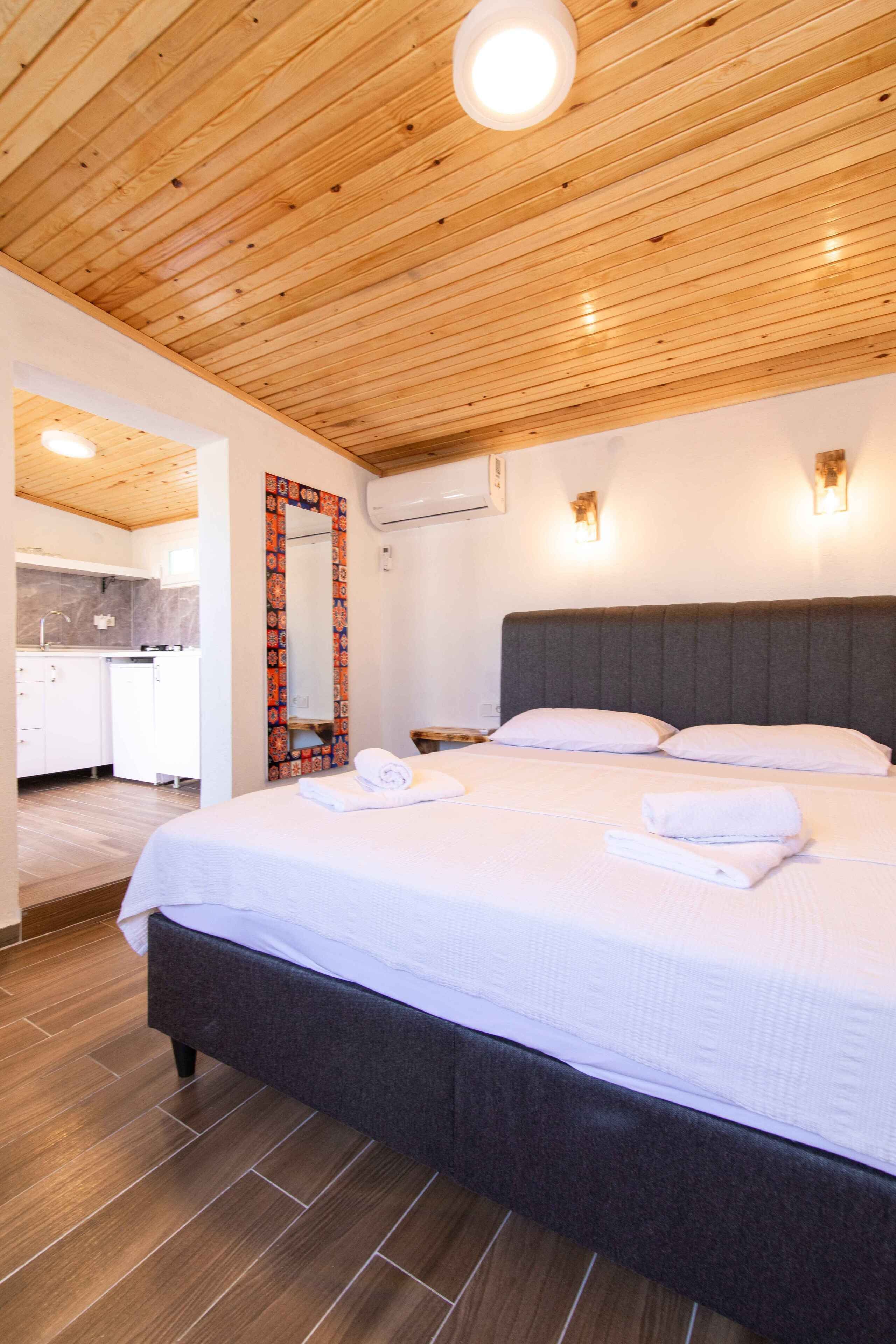 Una habitación de hotel moderna con techo de madera, una cama grande con ropa blanca, cabecero oscuro y un baño visible al fondo.