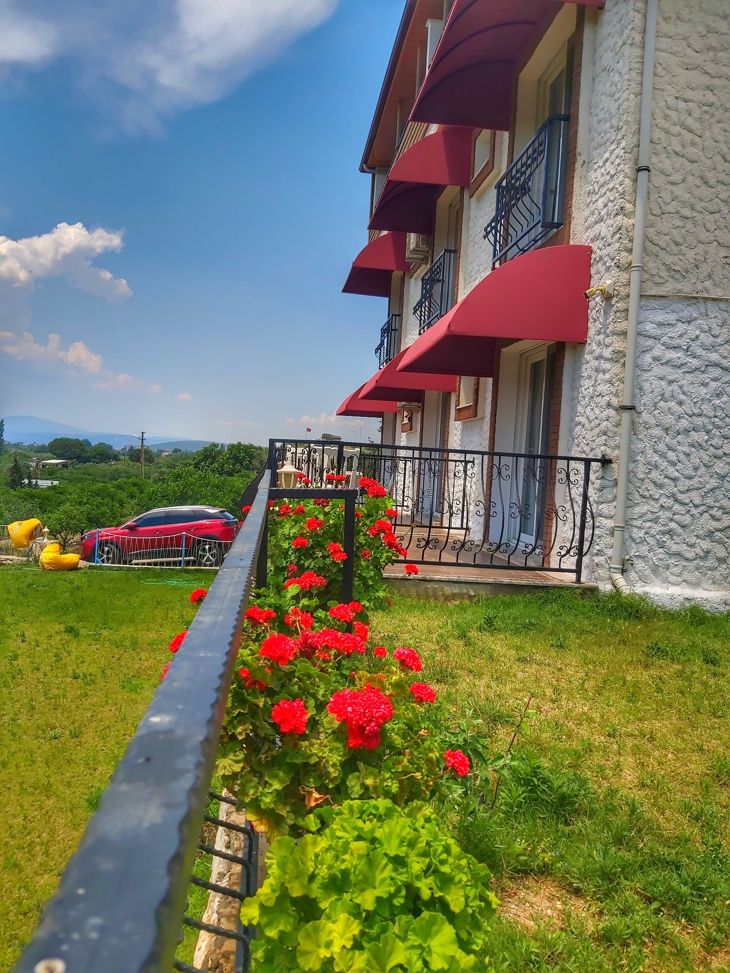 Un hotel con toldo rojo, un balcón con barandilla de metal y un jardín con flores rojas.