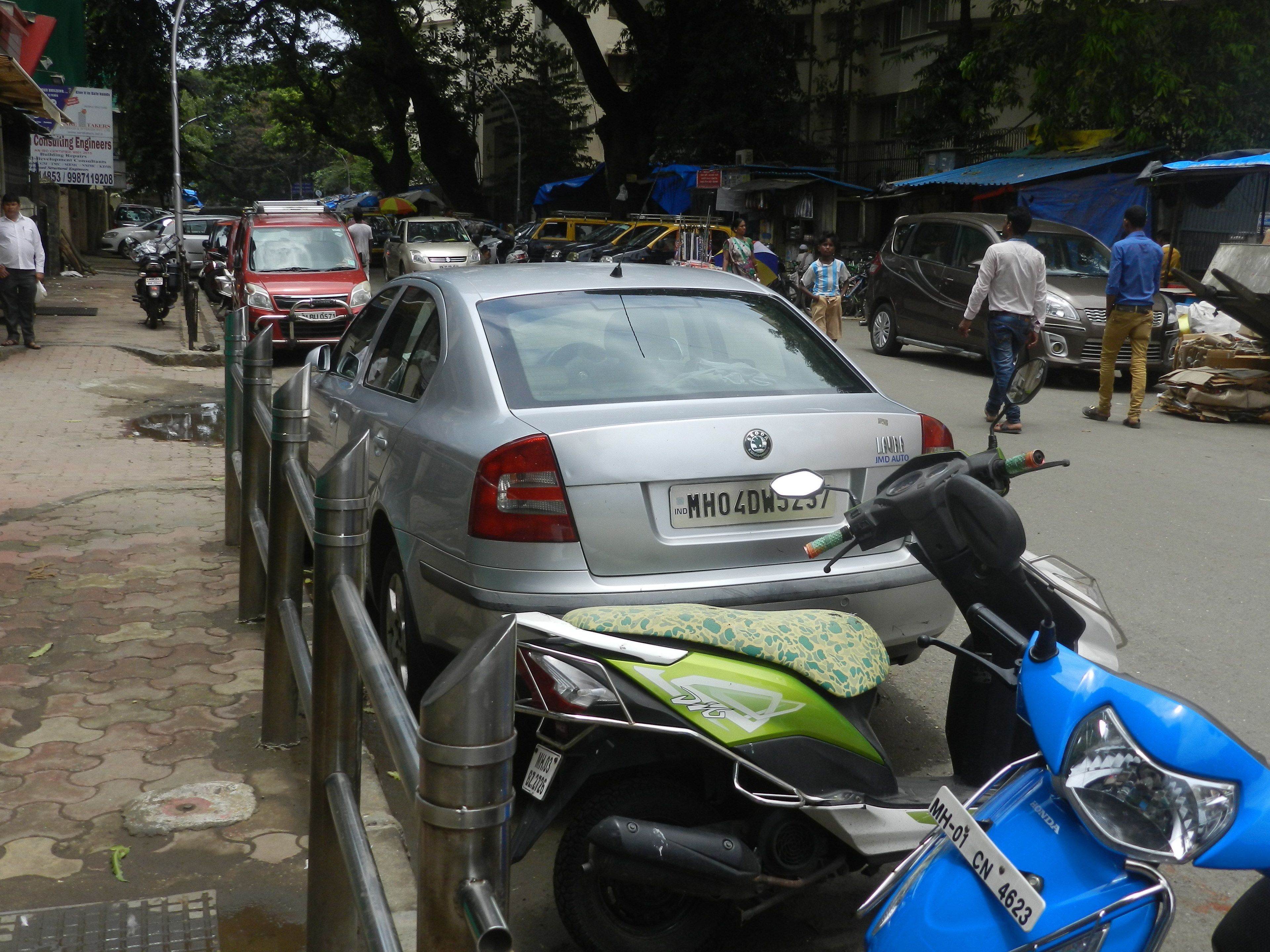 Escena callejera con coches y motocicletas estacionados, un paso de peatones y edificios al fondo.