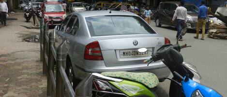 Escena callejera con coches y motocicletas estacionados, un paso de peatones y edificios al fondo.
