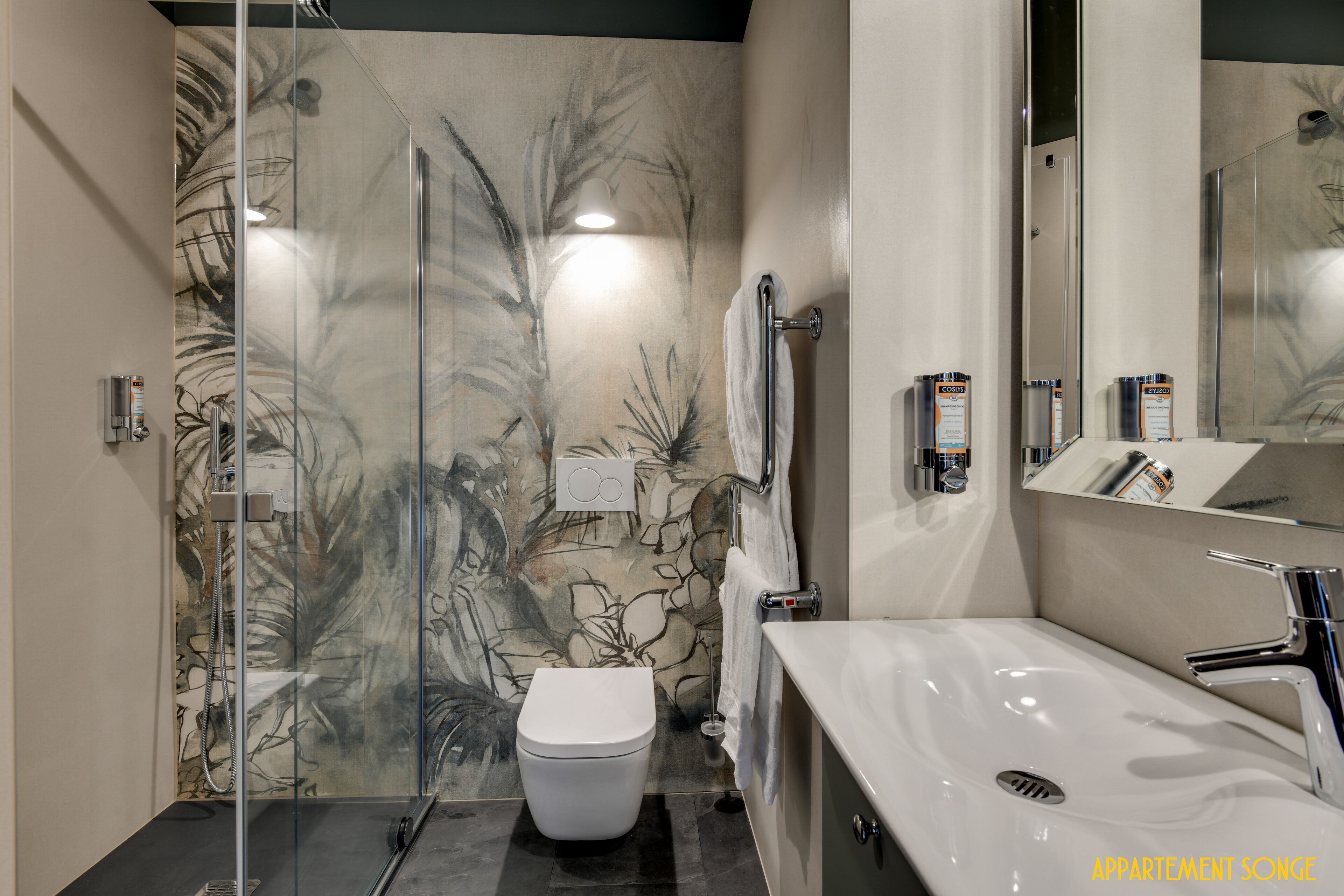 A modern bathroom with a glass shower enclosure, a white toilet, a sink with a mirror, and a wall with a leaf pattern.