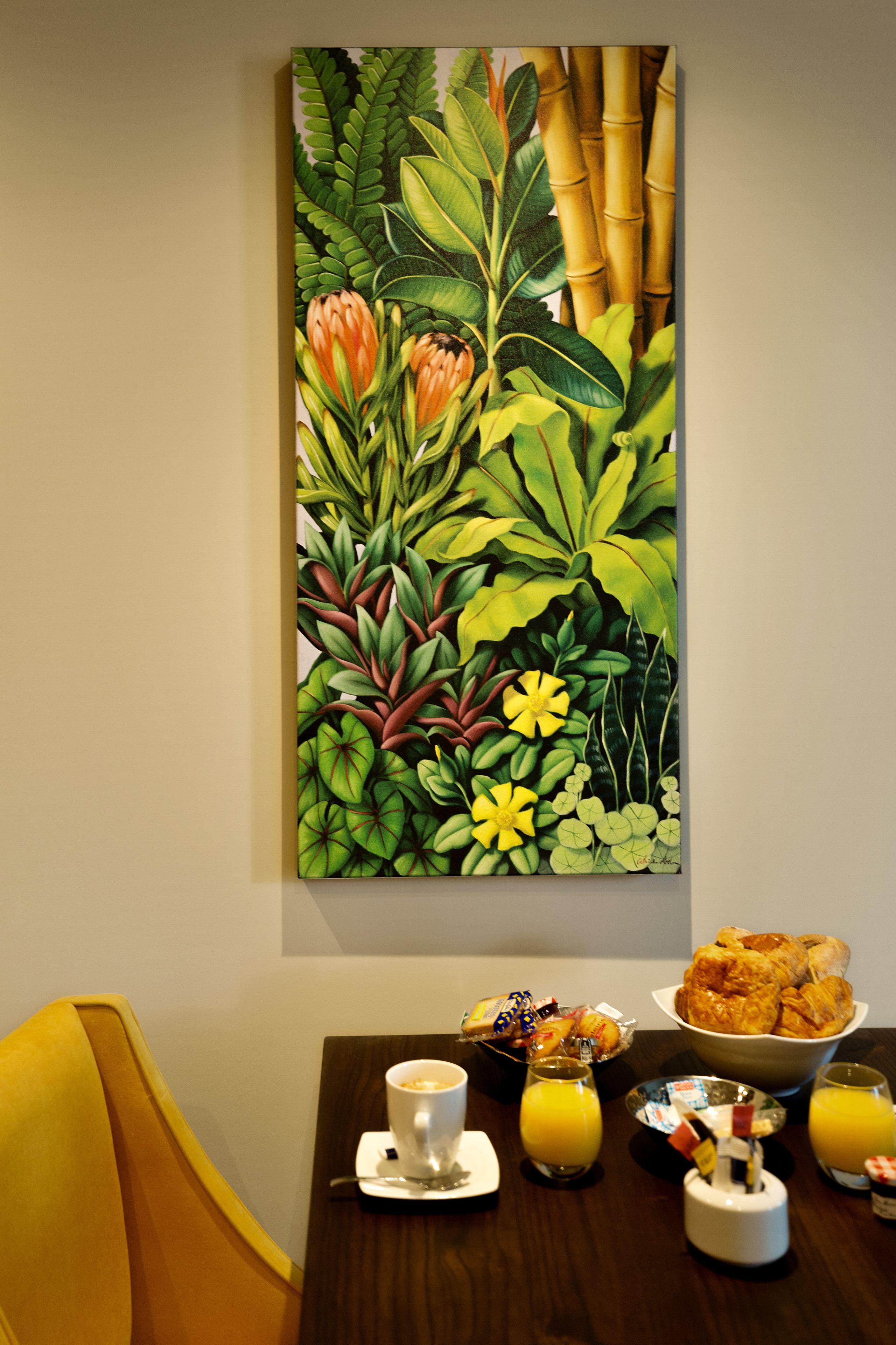 Une table à manger avec des aliments pour le petit-déjeuner, une tasse de café et un tableau représentant des plantes tropicales.