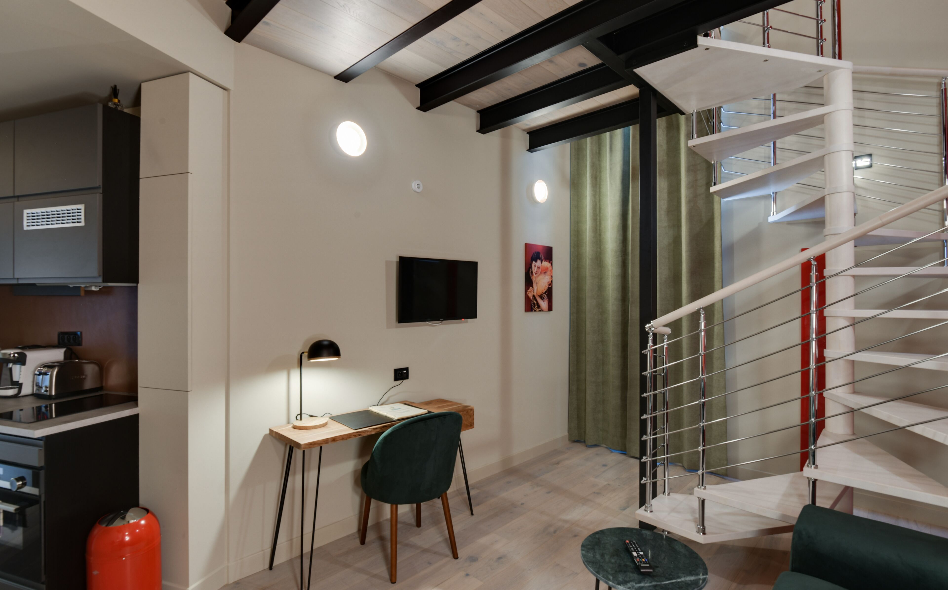 A modern kitchen with built-in appliances, a dining area with a desk and chair, and a staircase with a red railing.