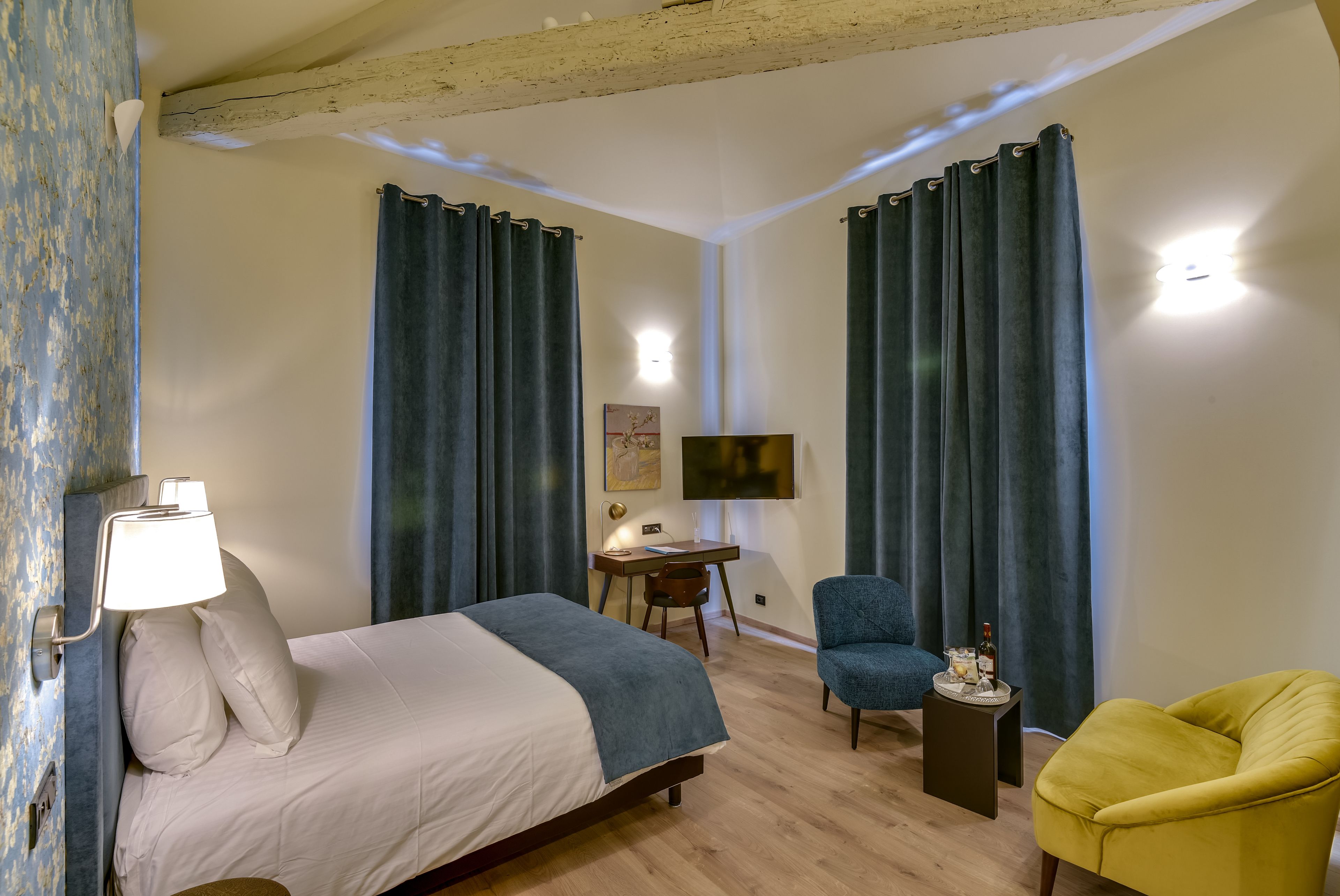 A hotel room with a bed, a desk, a chair, a TV, and a small table with a bottle and glasses.
