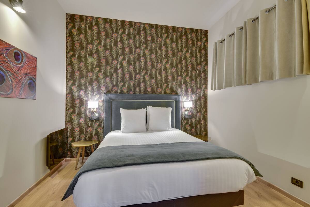 Une chambre d’hôtel avec un grand lit, une tête de lit, deux lampes fixées au mur, une table de chevet et un papier peint orné d’un motif de plumes de paon.