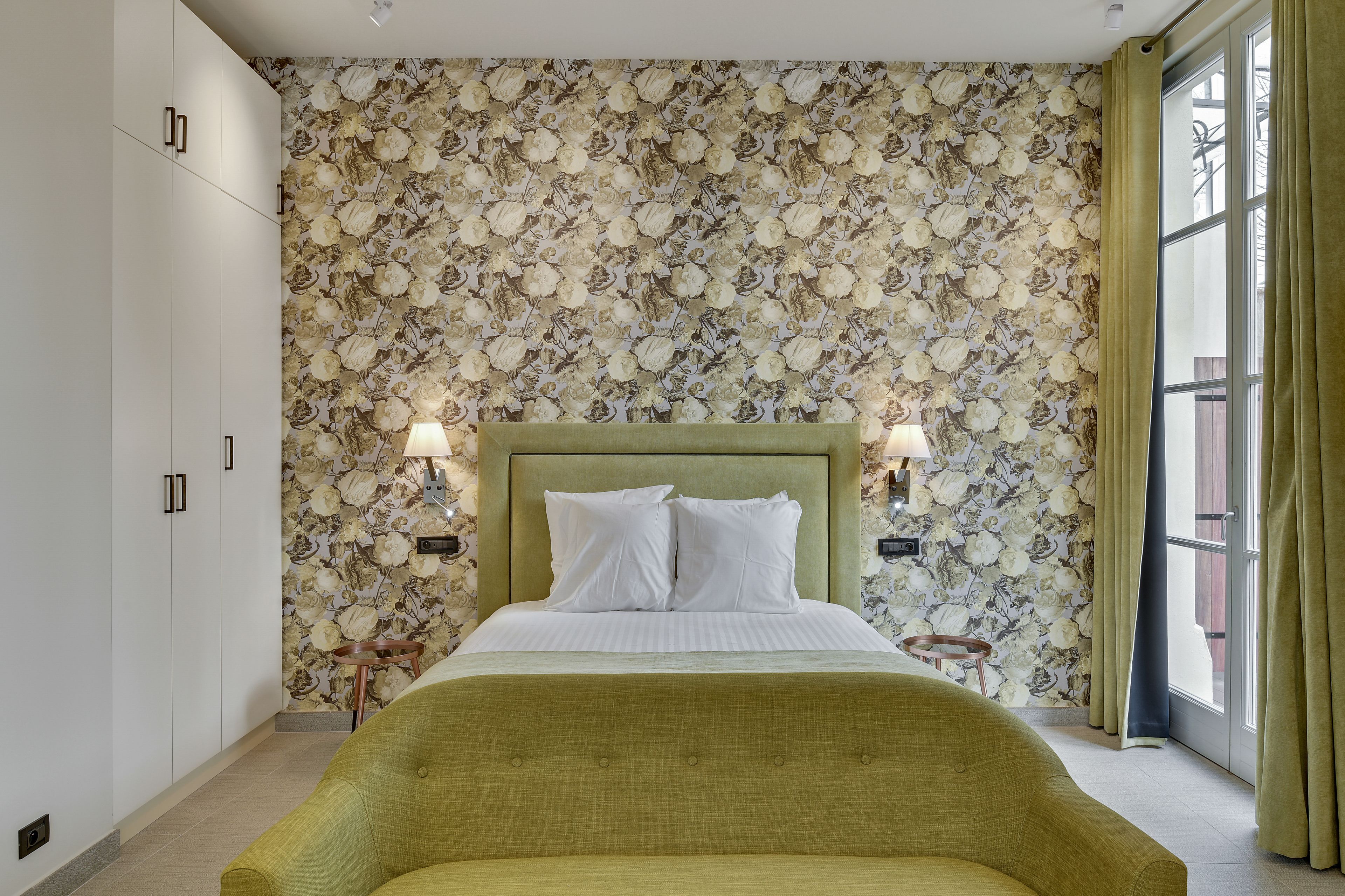 Un dormitorio con papel tapiz floral, un cabecero tapizado en verde, dos lámparas de noche y un ventanal con cortinas.