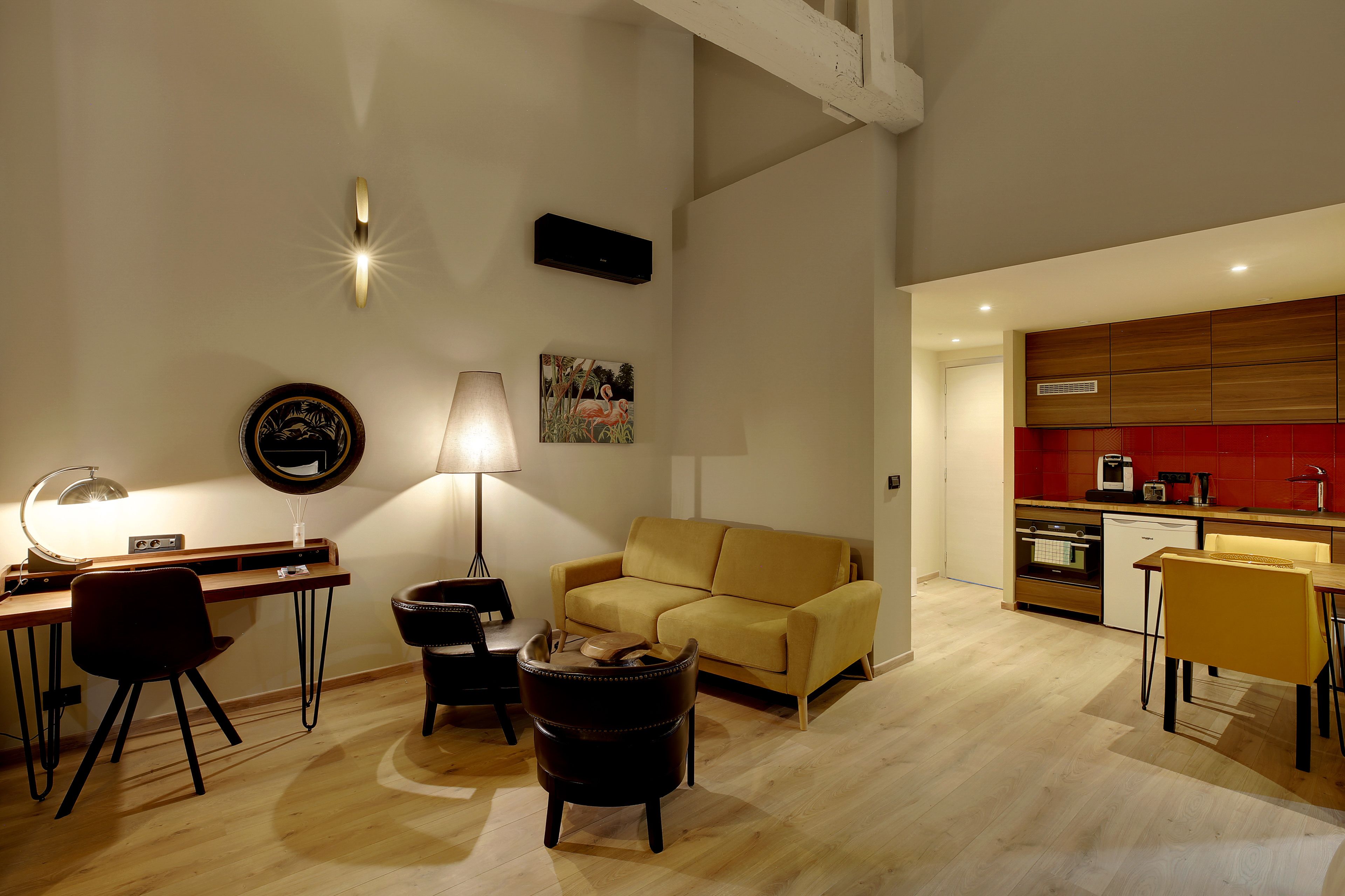 Ein modernes Wohnzimmer mit einer Couch, einem Schreibtisch und einer Lampe. Es gibt eine Küchenzeile mit roten Fliesen und Holzschränken.