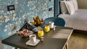 Une table de petit-déjeuner bien dressée, avec des croissants, du café et du jus d’orange, dans une chambre d’hôtel aux murs recouverts d’un papier peint à motifs floraux et dotée d’un lit avec du linge de maison blanc.
