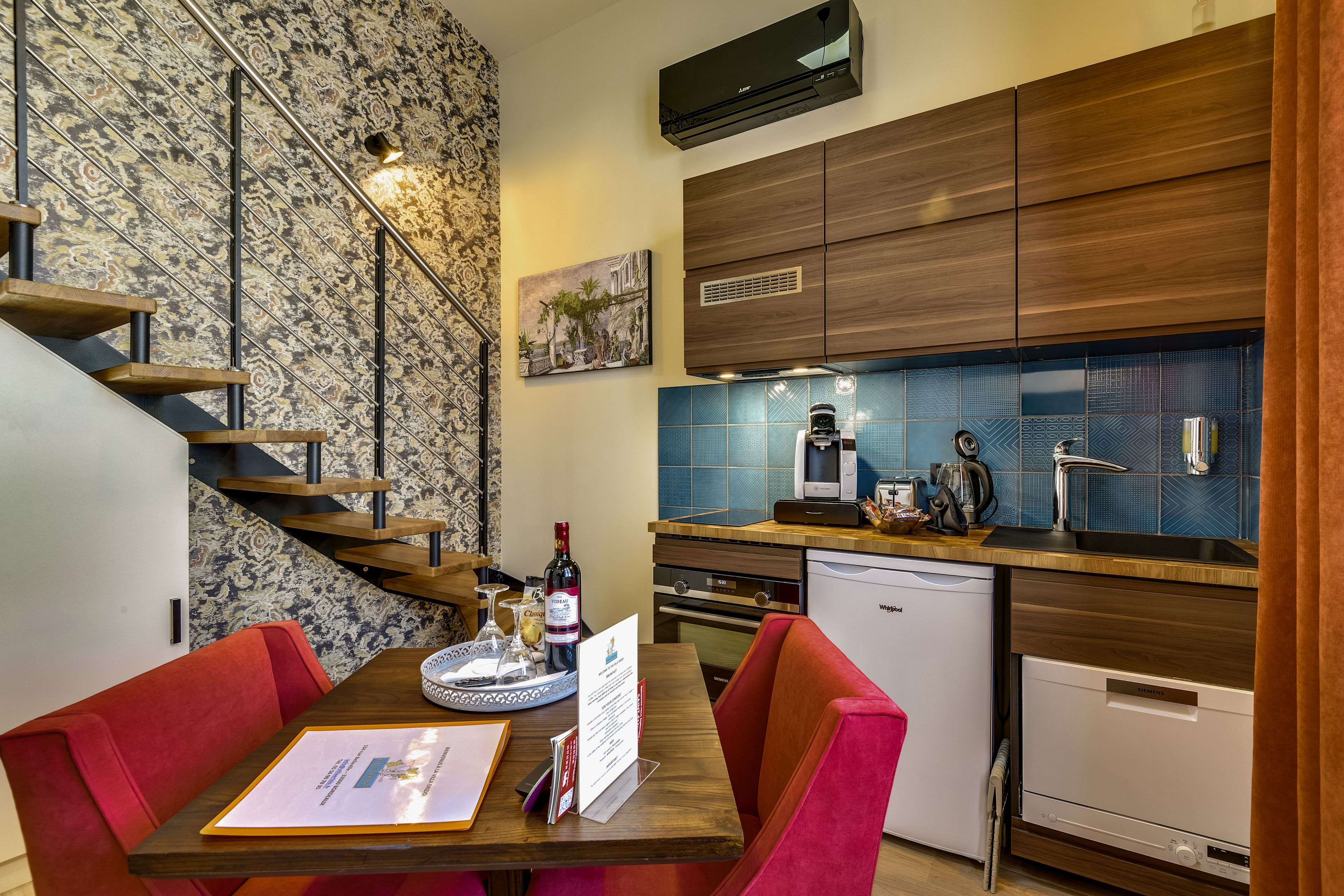 Een moderne keuken met houten kasten, een ingebouwde oven en een vaatwasser. Er is een eethoek met een tafel en stoelen, en een trap met een behang dat een patroon heeft.