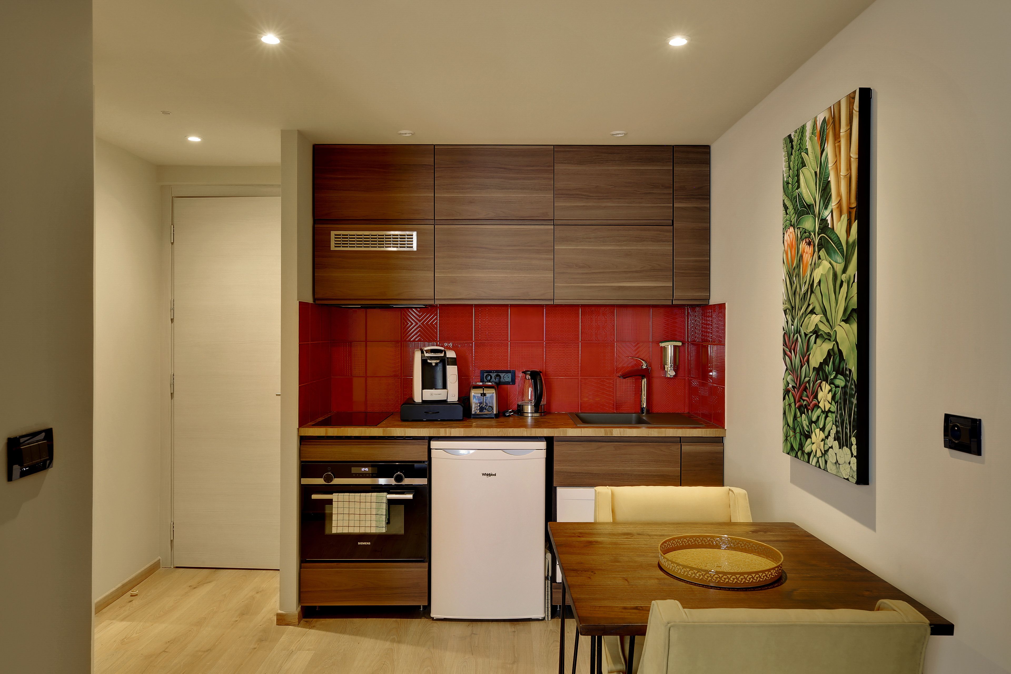 A compact kitchen with a refrigerator, microwave, and coffee maker. There is a dining table with chairs and a painting on the wall.