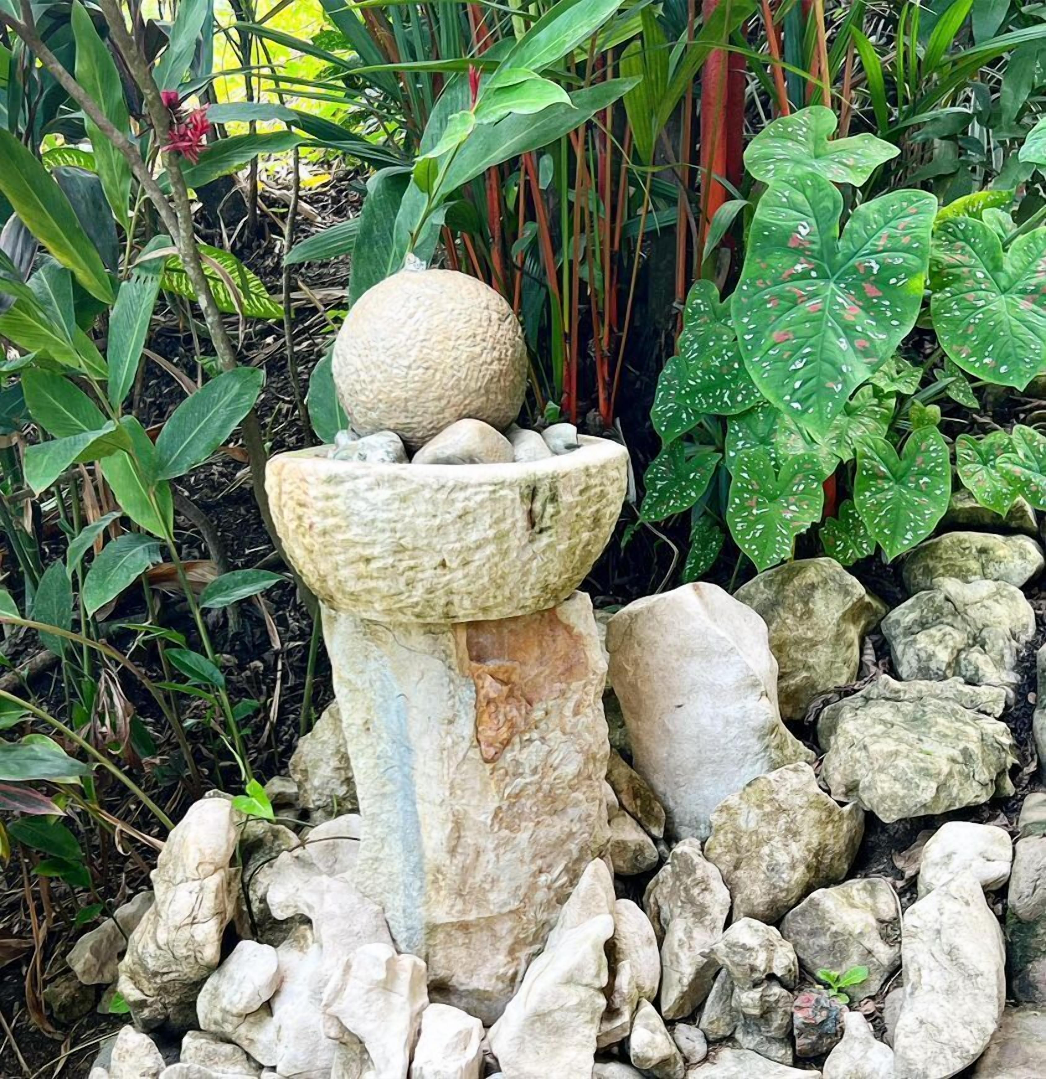 A garden fountain with a stone basin and a spherical stone on top, surrounded by rocks and greenery.
