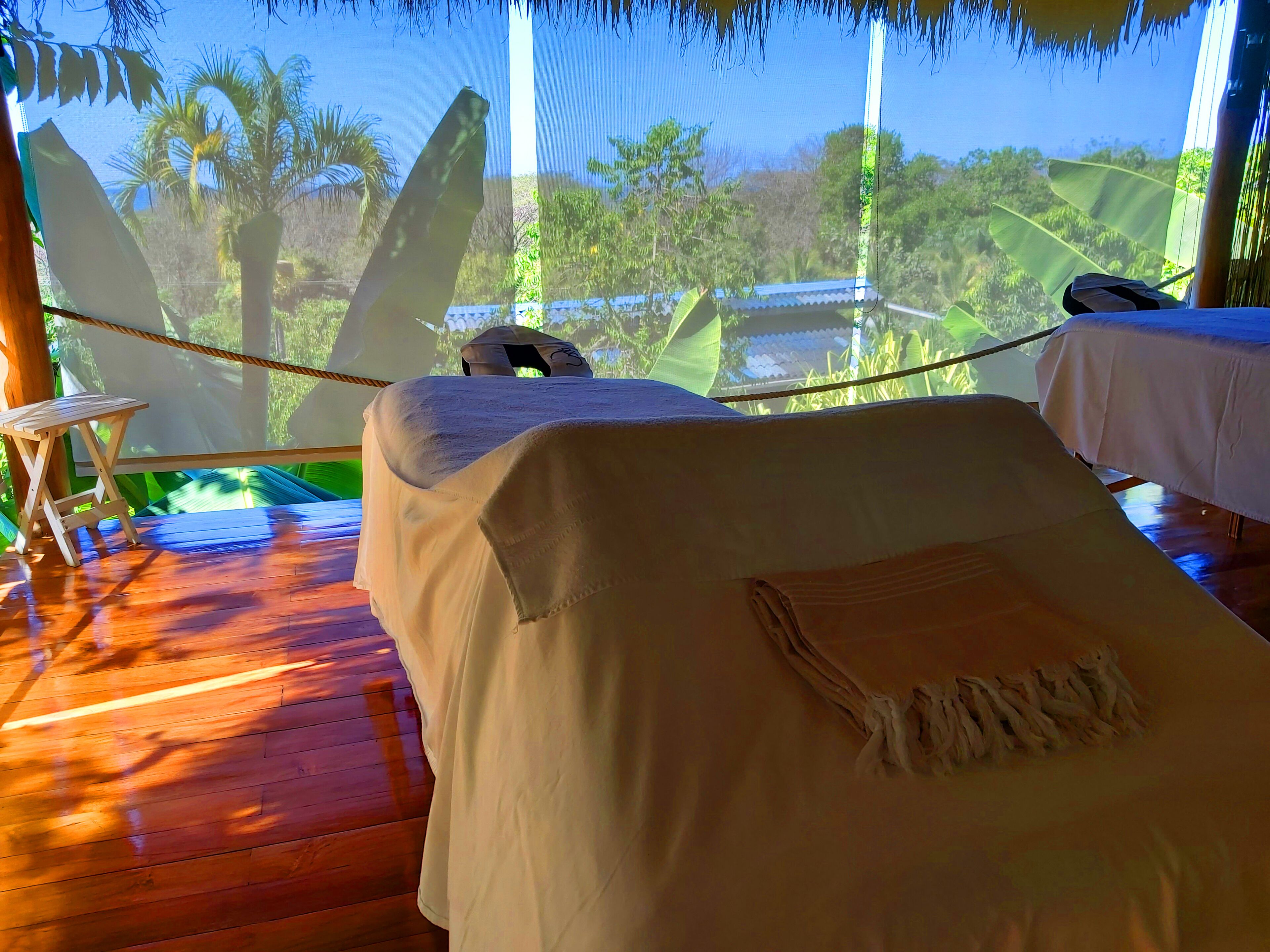 A room with two beds, a wooden floor, and a view of palm trees and a thatched roof structure.
