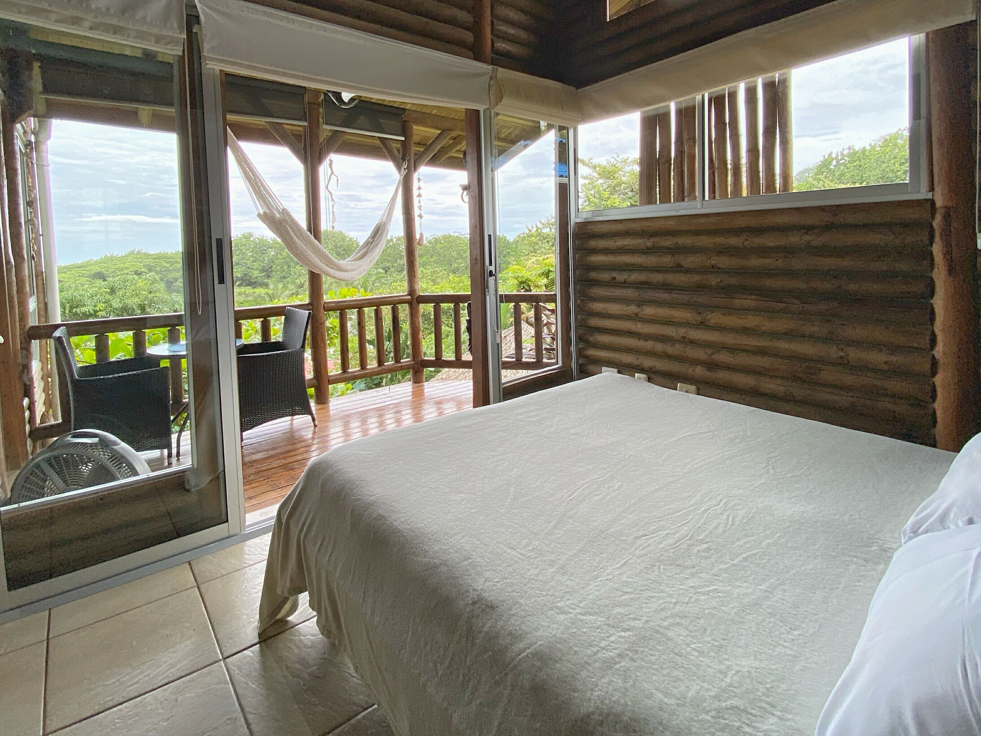 A bedroom with a large bed, a view of lush greenery, and a balcony with wicker furniture.