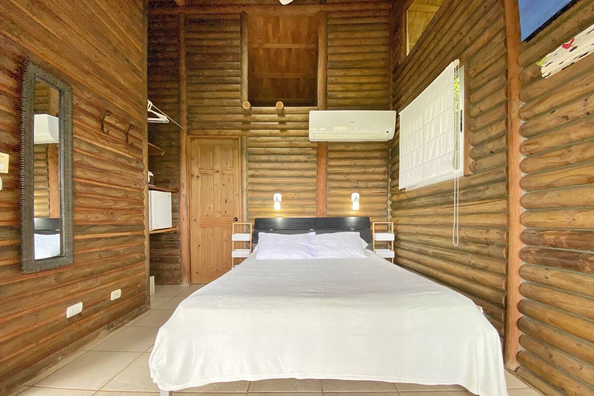 Une chambre située dans un chalet en rondins, avec un lit, un miroir, un climatiseur fixé au mur et une fenêtre avec des stores.