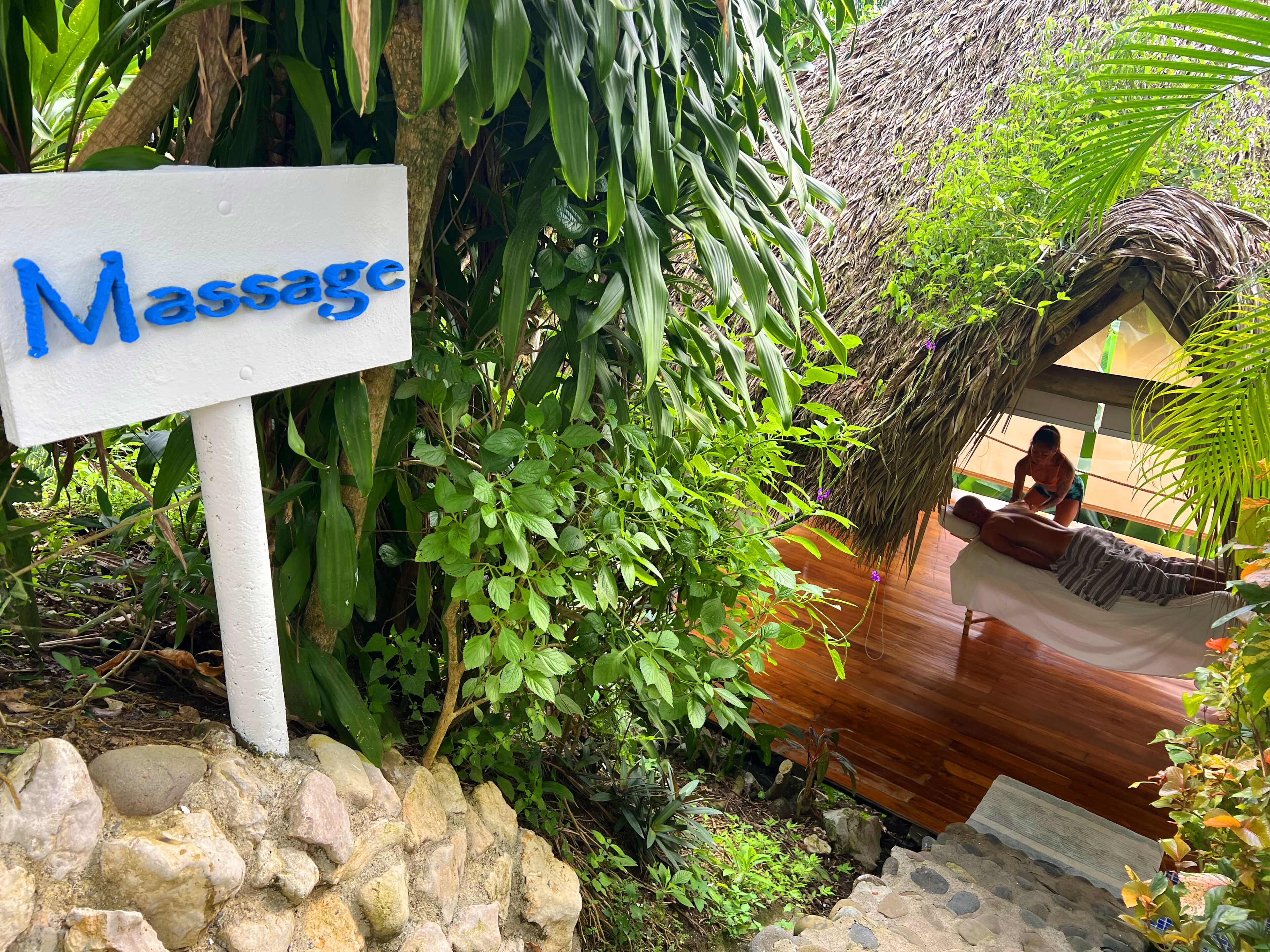 A massage area with a sign, surrounded by lush greenery and a thatched-roof structure.