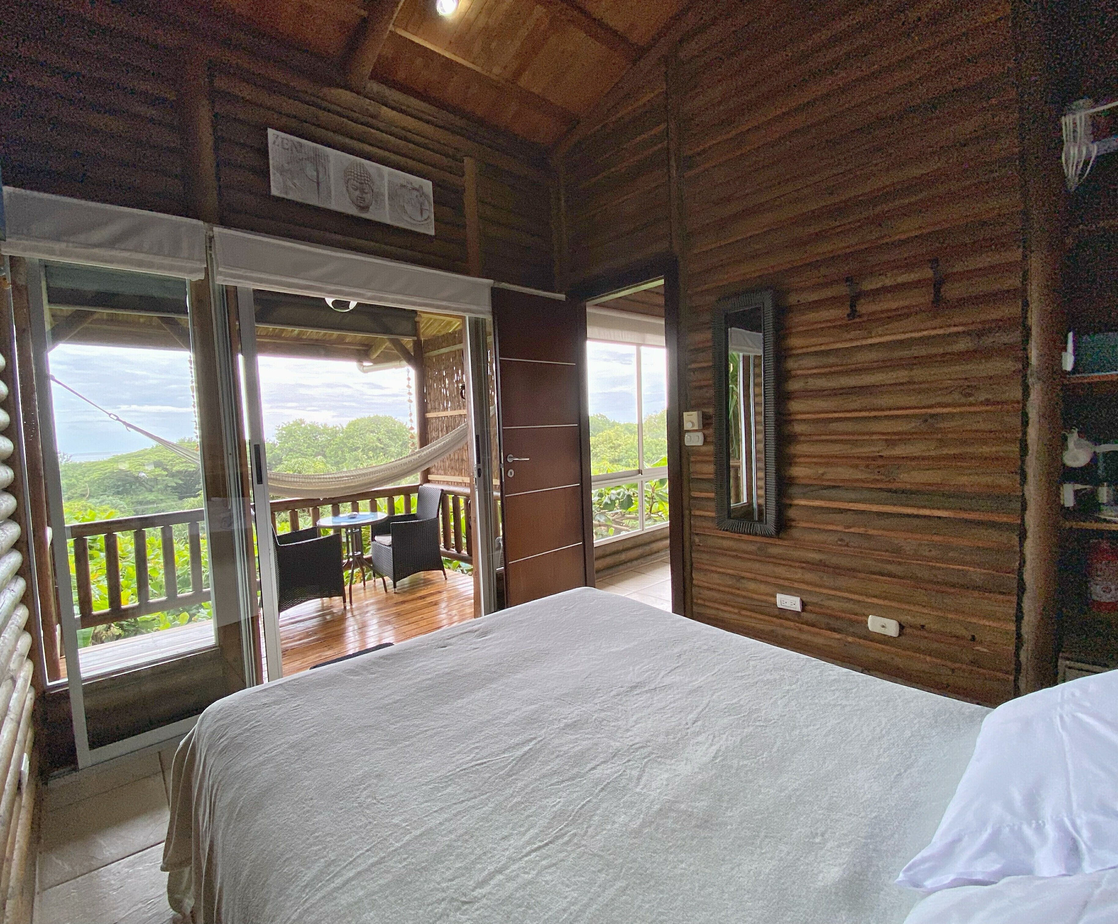 A bedroom with a large bed, a balcony with a table and chairs, and a view of greenery.