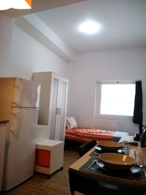 Cozinha compacta com geladeira, uma mesa pequena com tigela e utensílios, cama com manta listrada e janela com vista.