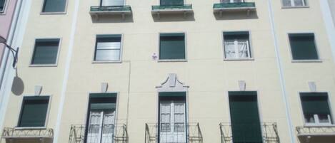 A multi-story building with a central entrance, balconies with metal railings, and multiple windows with shutters.