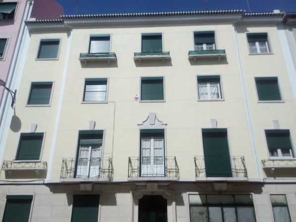 A multi-story building with a central entrance, balconies with metal railings, and multiple windows with shutters.