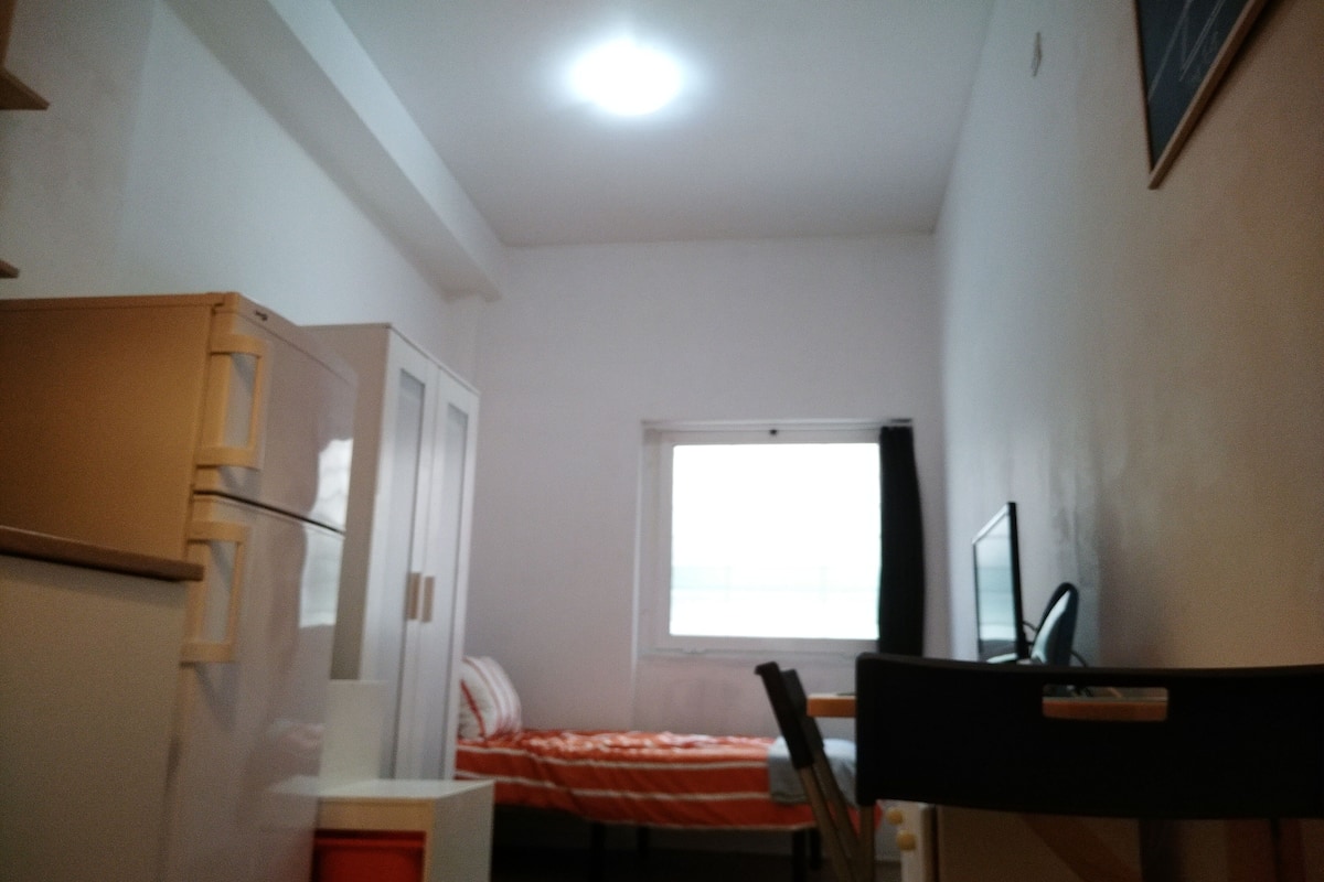 Een kleine, smalle kamer met een bed, een bureau en een stoel. Er is een raam met uitzicht op buiten.
