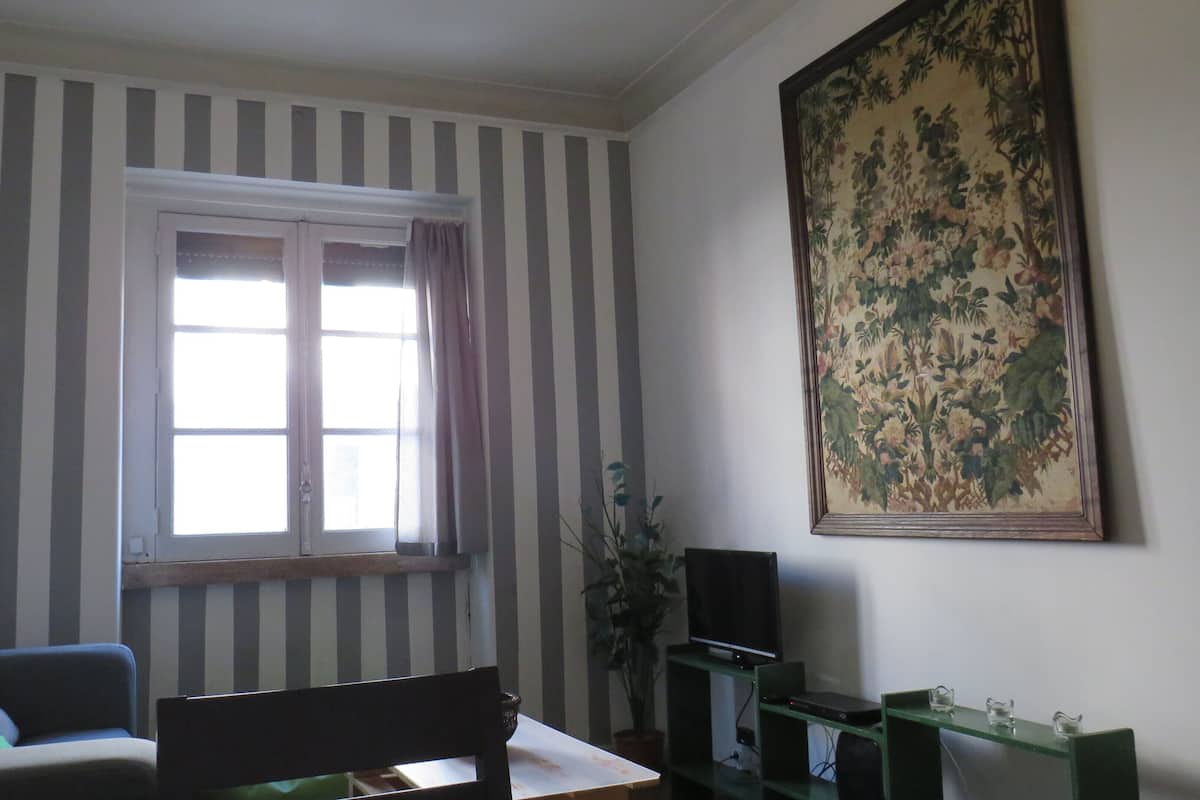 Een kamer met gestreept behang, een raam met gordijnen, een schilderij aan de muur, een televisietoestel en een tafel met een stoel.