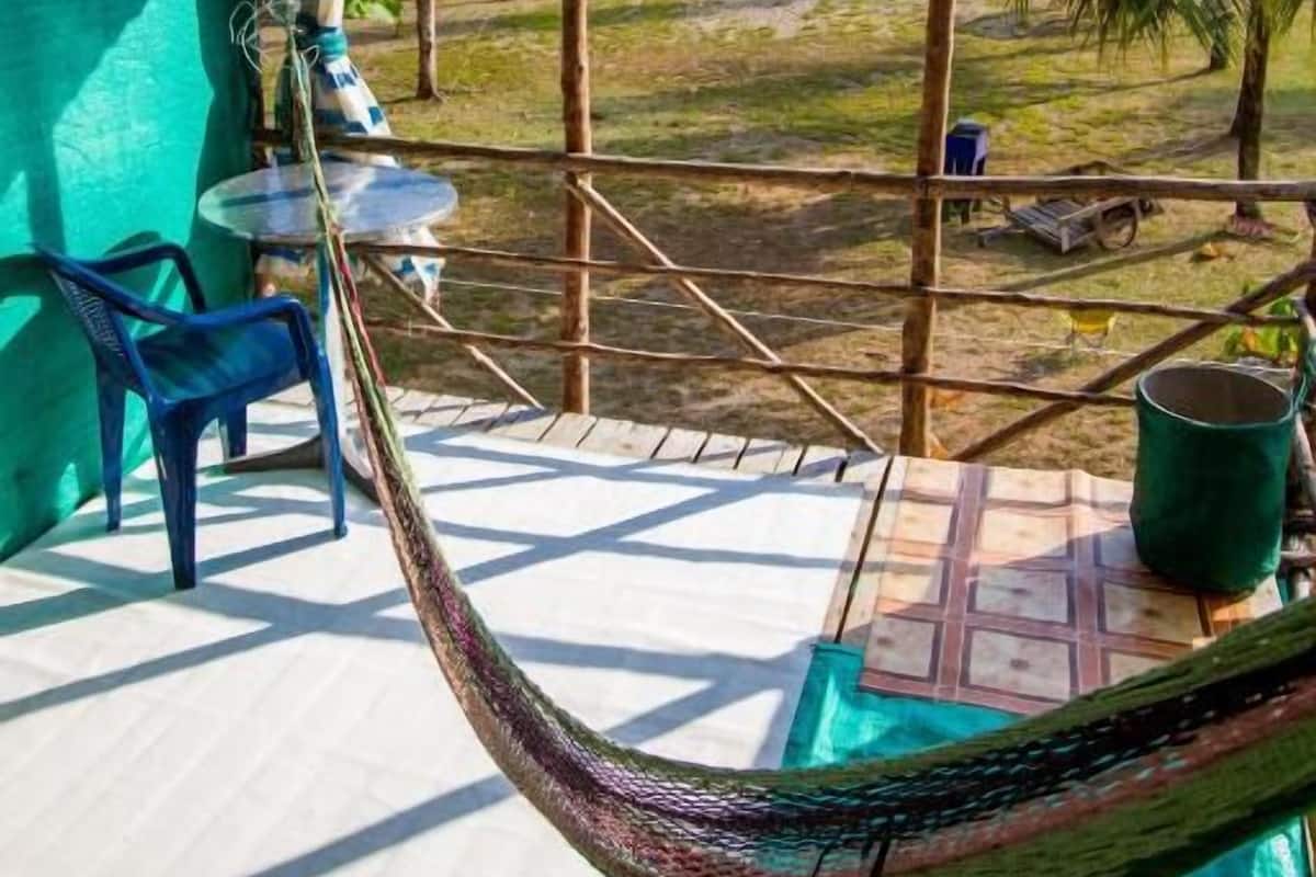 Una silla hamaca bajo un techo de lámina corrugada, una mesita y vista a una playa con palmeras.