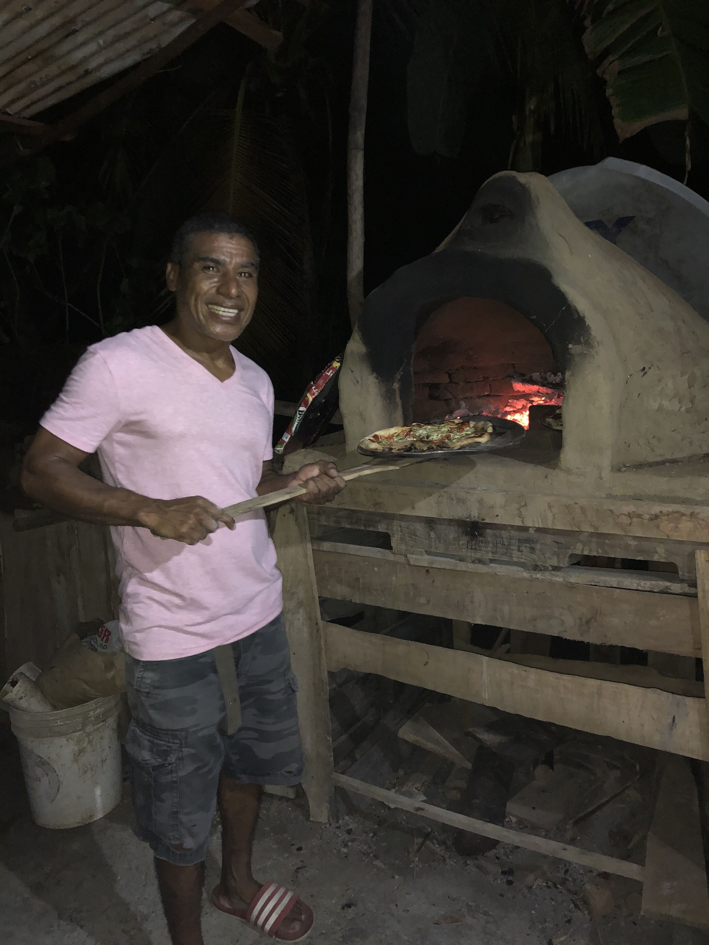 Une personne portant un t-shirt rose et un short de camouflage, tenant une pizza devant un four à bois.
