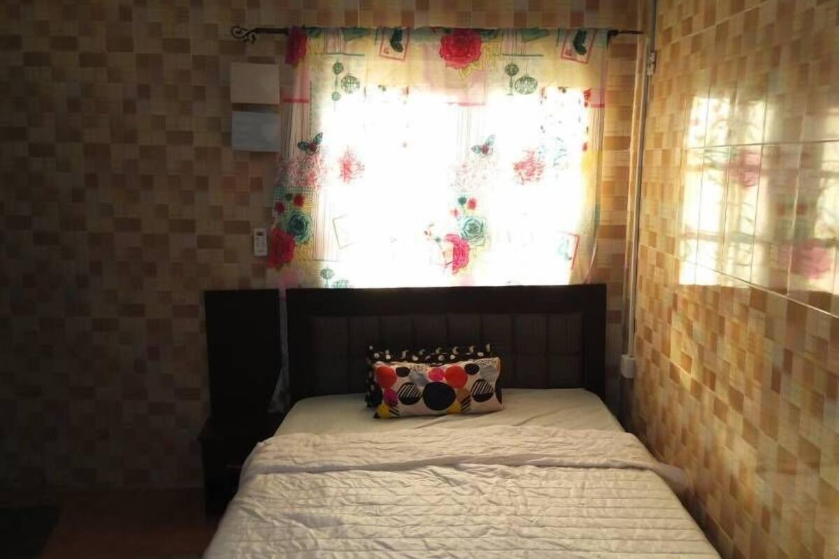 A bedroom with a bed, a pillow with a colorful pattern, a window with floral curtains, and an air conditioning unit on the wall.