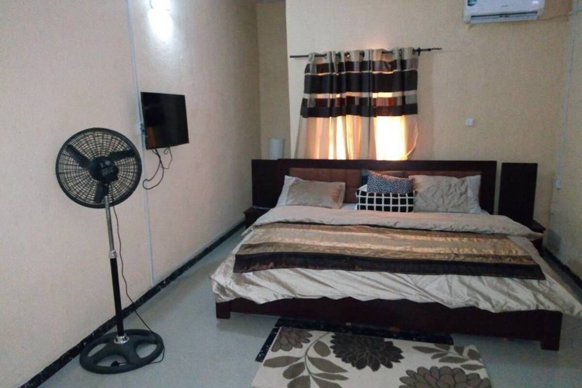 A bedroom with a bed, a standing fan, a wall-mounted TV, and a window with curtains.
