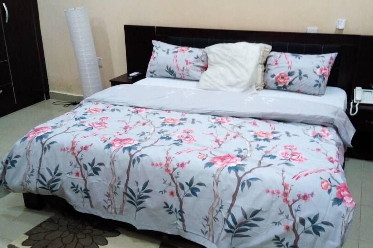 A neatly made bed with floral bedding, a dark headboard, and a floral-patterned rug on a tiled floor.