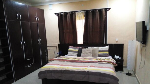 A bedroom with a bed, dark wooden wardrobe, curtains, a television, and a bedside table. - DABOTOV Hotel and Suites (Ilorin)