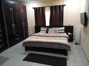 Quarto com cama, guarda-roupa de madeira escura, cortinas, televisão e mesa de cabeceira.