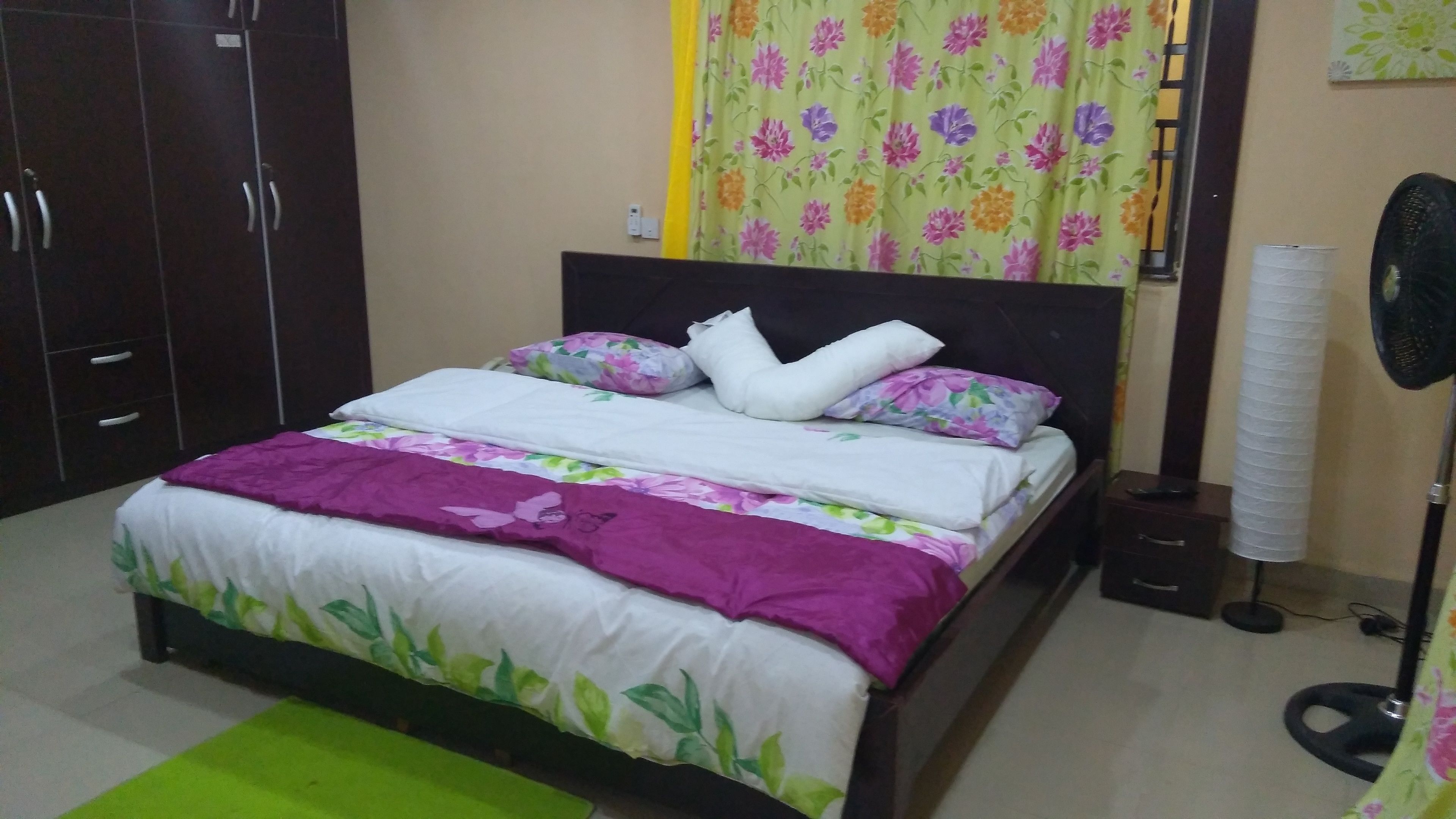Una cama bien hecha con ropa de cama floral, una alfombra verde y un ventilador de pie.