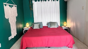 Ett sovrum med ett rött sängöverkast, vita gardiner och två sänglampor.
