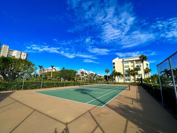 An outdoor tennis court surrounded by greenery and buildings.