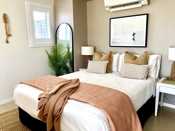 Un dormitorio con cama, mesitas de noche, un espejo, una planta en maceta y un cuadro en la pared.