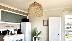 Una cocina compacta con un refrigerador blanco, una cafetera y un microondas encima. Hay una mesa de vidrio con sillas, una planta en maceta y una lámpara colgante de mimbre.