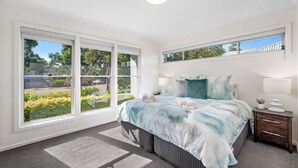 A bedroom with a large window, a bed with a blue and white bedspread, a bedside table with a lamp, and a small plant. - CYPRESS POINT LODGE - THE VINTAGE HOLIDAY HOUSES (POKOLBIN)