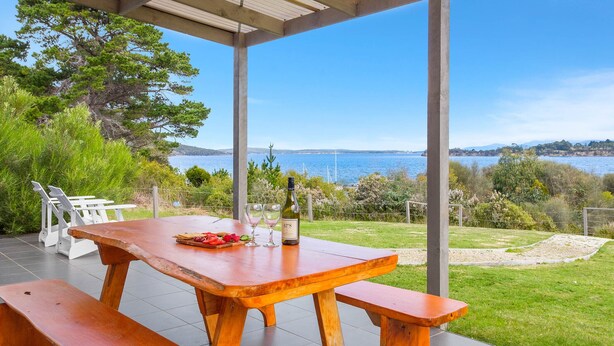 Une table en bois avec des bancs, une bouteille de vin et des verres, le tout sur une terrasse donnant sur un plan d’eau et une végétation luxuriante.