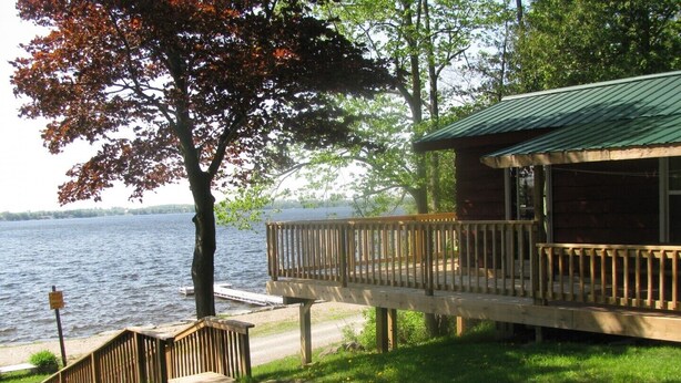 Una cabaña junto al lago con una terraza de madera y techo verde, con vista al agua.