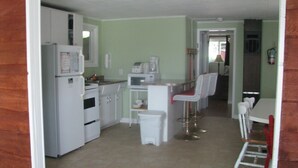 Cocina compacta con electrodomésticos blancos, un pequeño comedor con mesa y sillas, y un cubo de basura a la vista.