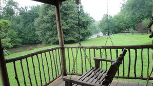 Une chaise suspendue en bois, installée sur une terrasse en bois, d’où l’on peut admirer une rivière et la verdure environnante.