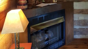 L’intérieur d’un chalet accueillant, avec une cheminée, des murs en bois, une lampe et une petite table sur laquelle sont posés des livres et des objets décoratifs.